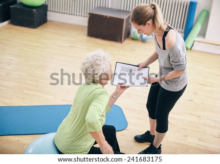 Senior woman in a gym sitting on exercise ball and talking to her private trainer about exercise plan