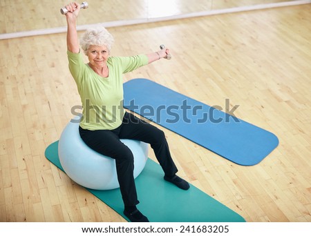 Active senior woman sitting on a pilates ball and lifting dumbbells looking at camera. Old caucasian woman exercising with weights at gym.