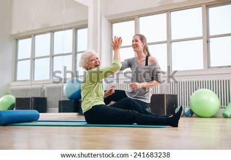 Senior woman sitting on fitness mat giving high five to her personal trainer at gym. Excited old woman rejoicing health success with her instructor at rehab.