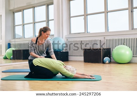 Female trainer helping senior woman doing yoga. Elder woman bending over a exercise mat with personal instructor helping at gym.