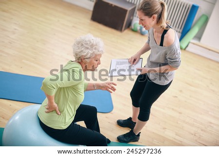 Senior woman sitting on a fitness ball with her female instructor explaining exercise plan at gym. Physical therapist with old woman at rehab.
