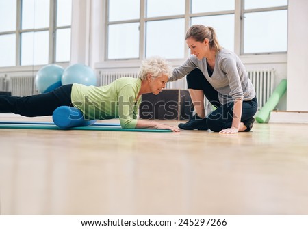 Senior woman exercising with a foam roller being assisted by personal instructor at gym. Physical therapist helping elderly woman in her workout at health club.