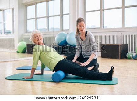 Senior woman doing pilates on the floor with foam roller. Elder woman exercising being assisted by personal trainer at gym.