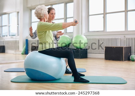 Female trainer assisting senior woman lifting weights in gym. Senior woman sitting on pilates ball doing weight exercise being assisted by personal trainer at health club.