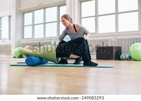 Female trainer instructing senior woman going exercise on a foam roller. Elder woman doing pilates workout with personal instructor at gym.