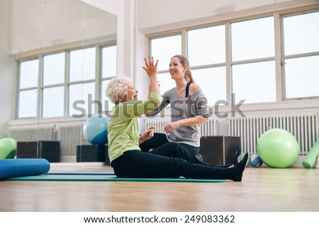 Senior woman giving high five to her personal trainer while sitting on fitness mat at gym. Happy elder woman rejoicing health success with her trainer.