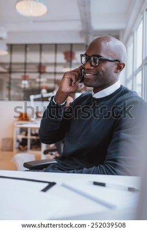 Happy young african man sitting at his desk talking on his mobile phone in office. African executive using cell phone while at work.