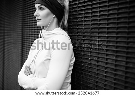 Black and white image of young woman wearing earphones standing leaning a wall looking away confidently. Fitness female taking a break for training session.