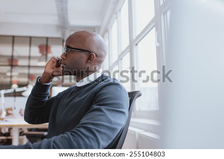 Side view of african executive sitting at his desk using mobile phone. Young man at work answering a phone call.