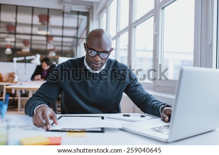 Young african executive sitting at his desk with laptop reading a document. African man working in office.