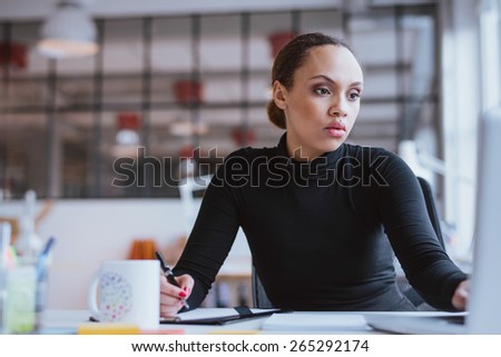 Image of african young woman working new business assignment. Female executive sitting at her desk using laptop and writing notes at office.