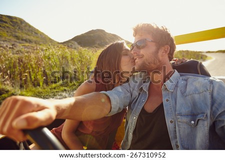 Woman kissing her boyfriend driving a car. Lovely young couple on road trip. Affectionate caucasian couple enjoying road trip.