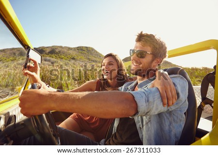 Man driving beach buggy with woman taking selfie on her smart phone. Couple having fun on road trip on a summer day.