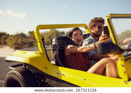 Excited happy couple enjoying road trip in their open top car. Man driving the car young woman taking a selfie from her mobile phone.