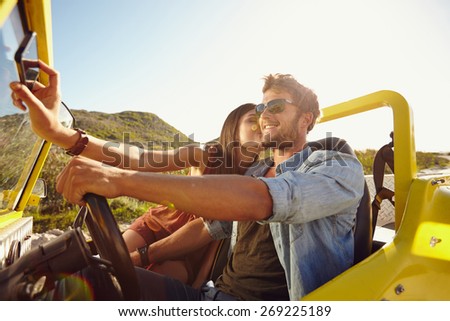 Woman kissing man and taking selfie on her mobile phone. Couple on road trip, man driving a car on a summer day.