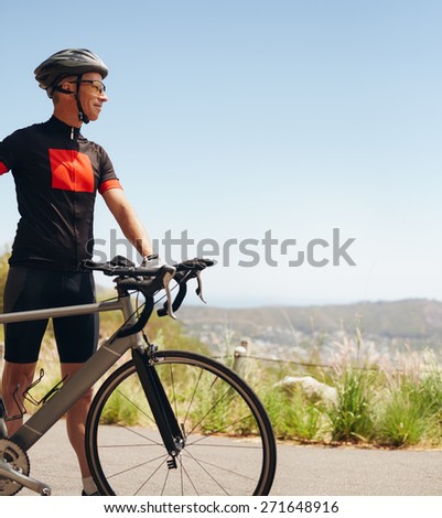 Image of happy male cyclist with his bike on the country side. Young cyclist standing with his bicycle looking away, outdoors.