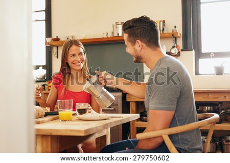 Indoor shot of young man serving coffee to his girlfriend having breakfast in kitchen at home. Smiling young couple having breakfast and looking at each other.