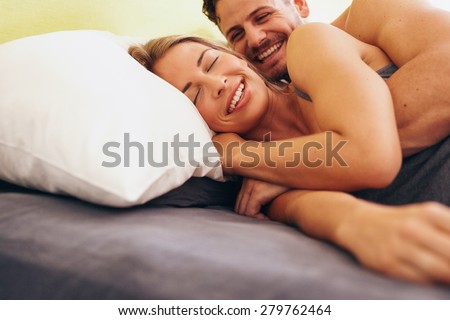 Happy young couple embracing while lying next to each other on bed. Caucasian couple smiling in bed together. Couple waking up.
