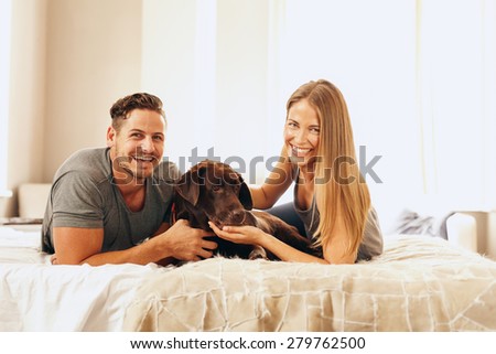 Shot of a young couple lying on the bed with their dog. Cheerful man and woman looking at camera with their pet in bedroom.