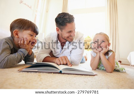 Portrait of happy young family of three lying on floor with a book. Father with two kids reading a story book in living room at home.