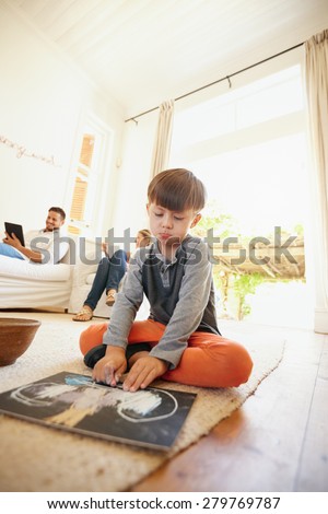 Little schoolboy sitting on floor drawing with his parents sitting in background on couch. Family in living room.