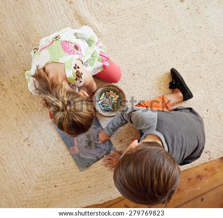 Top view of two little kids sitting on floor drawing with color chalks.