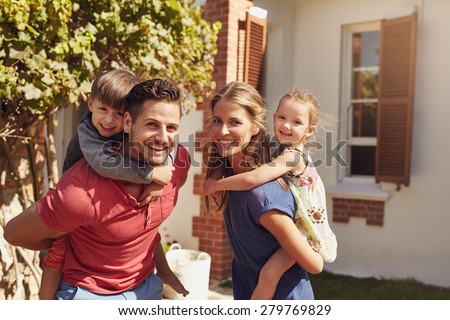 Happy father and mother looking at camera while piggybacking their son and daughter. Happy young family of four playing in their backyard on a sunny day.