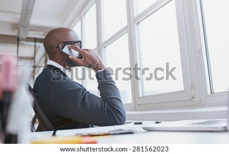 Young african man sitting at his desk talking on his mobile phone in office. African executive using cell phone while at work.