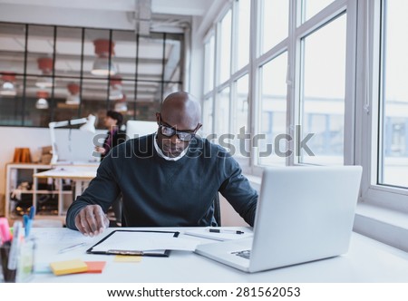 Young executive sitting at his desk with laptop reading a document. African man working in office.