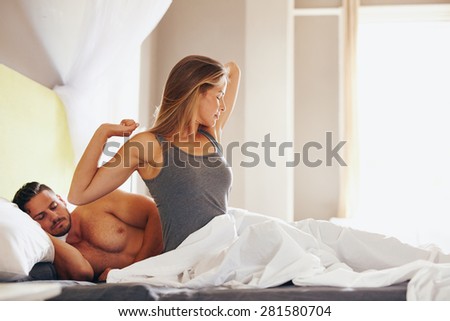Relaxed young woman stretching in bed with her husband sleeping behind her. Woman waking up in morning.