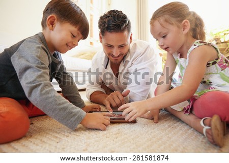 Father with son and daughter sitting on floor using digital tablet indoors. Happy young family together at home using touchpad computer. Young man teaching his children how to use digital tablet.