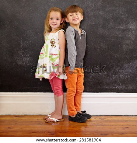 Little boy and girl standing back to back in front of blackboard looking at camera smiling. Full length image of two innocent little children together at home.