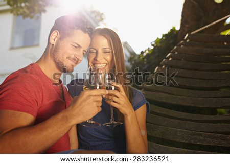 Outdoor shot of young caucasian couple in backyard toasting wine smiling. Romantic couple relaxing on hammock celebrating with wine.