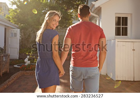 Young caucasian couple walking in the backyard holding hand in hand on a bright summer day, with woman looking back over shoulder and smiling at camera.