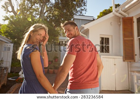 Rear view shot of a young couple taking a walk around their house holding hands. Loving young couple outdoors in their backyard on a bright sunny day looking back at camera smiling.