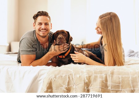 Portrait of happy young couple on bed with dog in morning. Man stroking the dog and smiling.