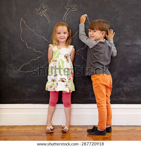 Full length portrait of cute little girl standing and little boy drawing angel wings around her on blackboard. Two little kids standing in front of blackboard at home looking at camera smiling.