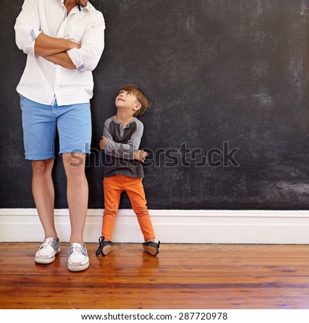 Indoor shot of little boy and his father standing with hands folded against black wall. Father and son looking at each other with copy space. Son imitating his father\'s pose.