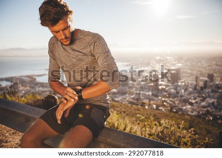 Young man adjusting his GPS watch before a run. Fit young athlete sitting on road railing and checking his watch while out for a run in morning.
