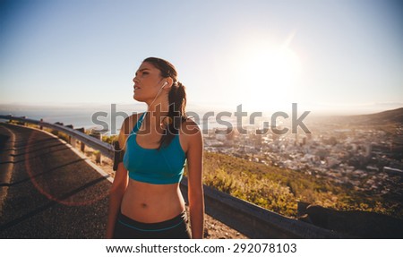 Image of young woman taking a break after hard training session on sunny day. Female athlete standing outdoors looking away.