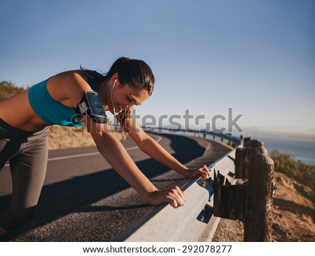 Tired runner taking a break leaning on country road guardrail. Fit young woman athlete training outdoors.
