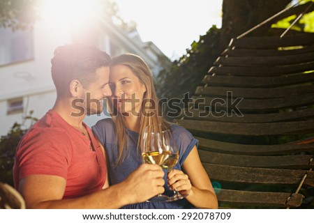 Romantic young couple sitting on garden hammock toasting wine. Loving young couple celebrating with a glass of wine in backyard on a bright sunny day.