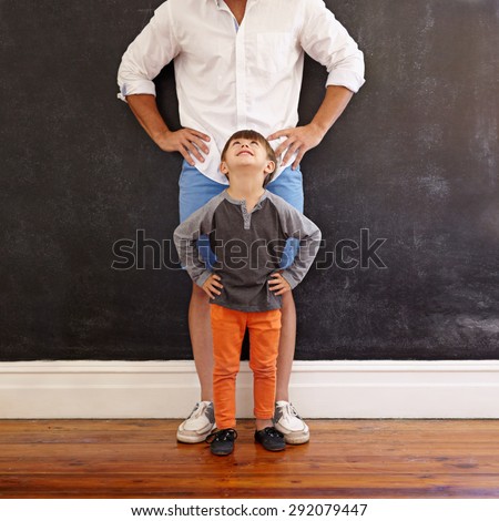 Indoor shot of little boy looking at his father while standing in front with hands on waist.