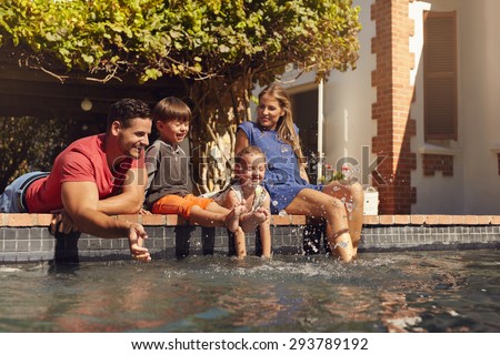 Outdoor shot of happy young family splashing water with hands and legs while sitting on edge of swimming pool. Family enjoying a hot sunny day playing by the pool.