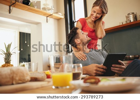 Happy young couple in their kitchen in morning. Man sitting at breakfast table with a digital table with woman standing next to him. Both looking at each other smiling.