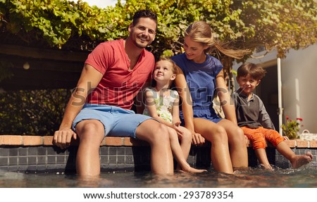 Outdoor shot of beautiful young family sitting next to swimming pool with their feet in water. Young couple with their kids enjoying a hot summer day near pool.