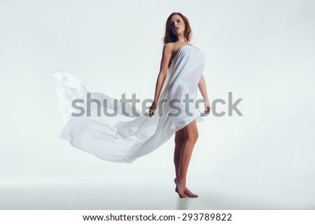 Sensual young woman posing with a white scarf on her body. Naked woman in transparent cloth looking away while standing over white background.