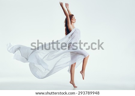 Side view of beautiful naked woman with flying white cloth on her body standing on one leg and hands raised. Sensual model posing over white background with copyspace.