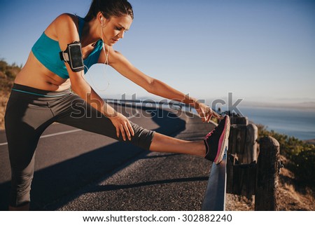 Female runner stretching her legs outdoor before running. Woman doing leg stretch exercises on road guardrail.