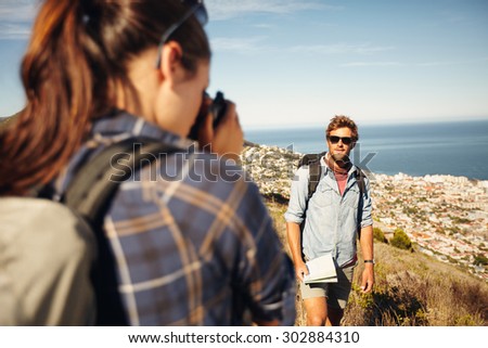 Young man posing in nature, with woman talking his pictures in countryside on a summer day while hiking. Young woman taking photo for her boyfriend with a camera.